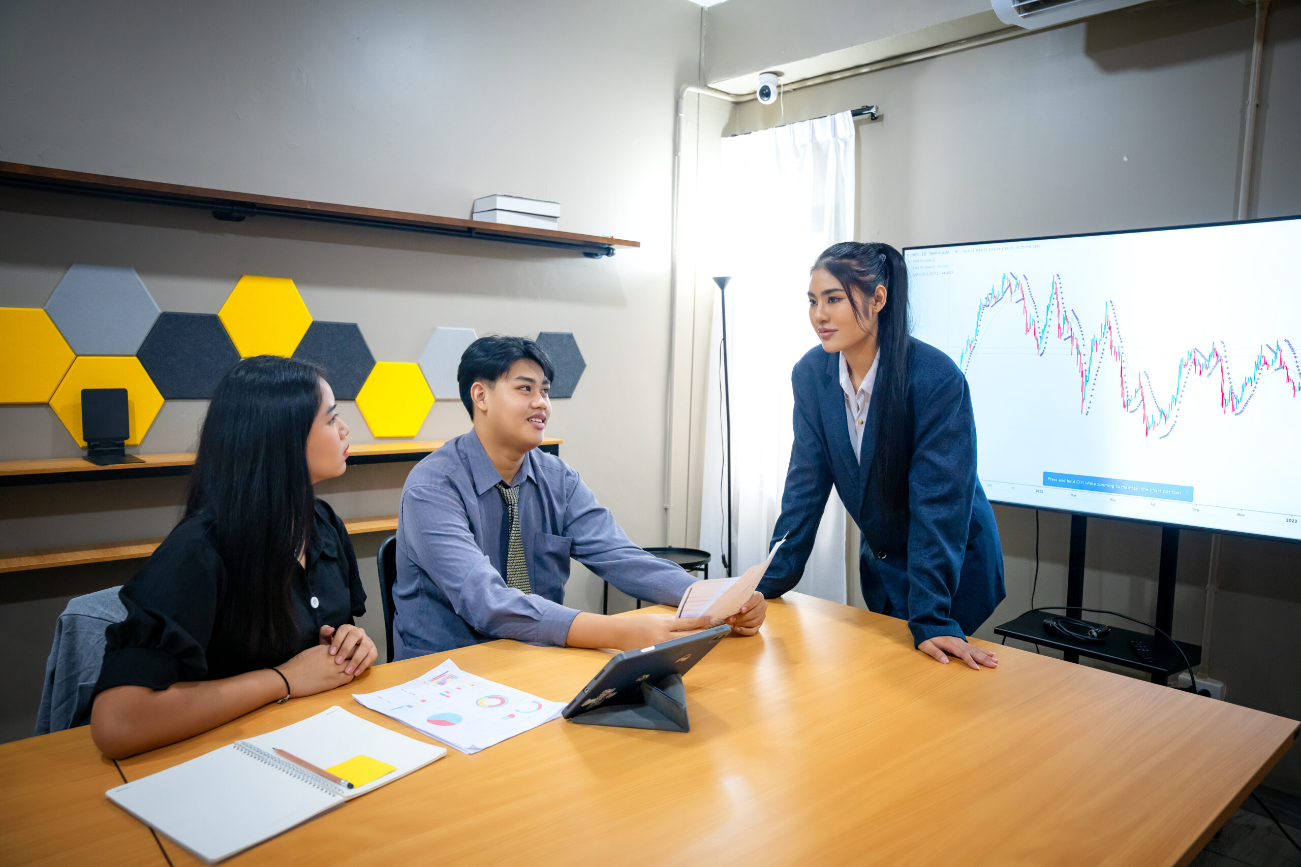 Three professionals gather in a contemporary office to analyze financial data displayed on a screen. They engage in discussion, highlighting graphs and trends to inform business decisions.