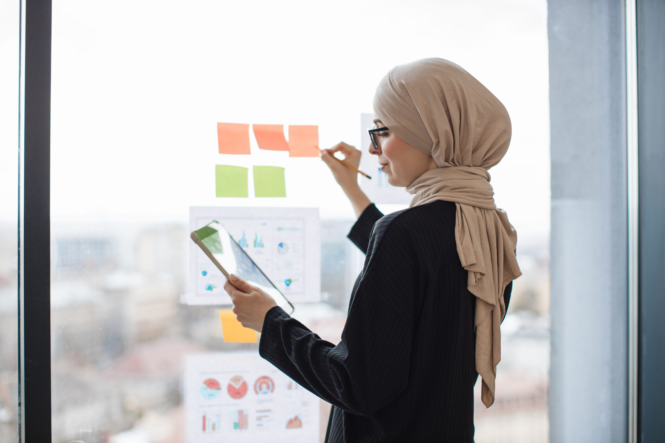 Lady with tablet writing notes on stickers near infographics Side view of arabian office administrator with tablet writing brief notes on stickers attached to window glass. Stylish entrepreneur in hijab analysing accounting records via infographics.
