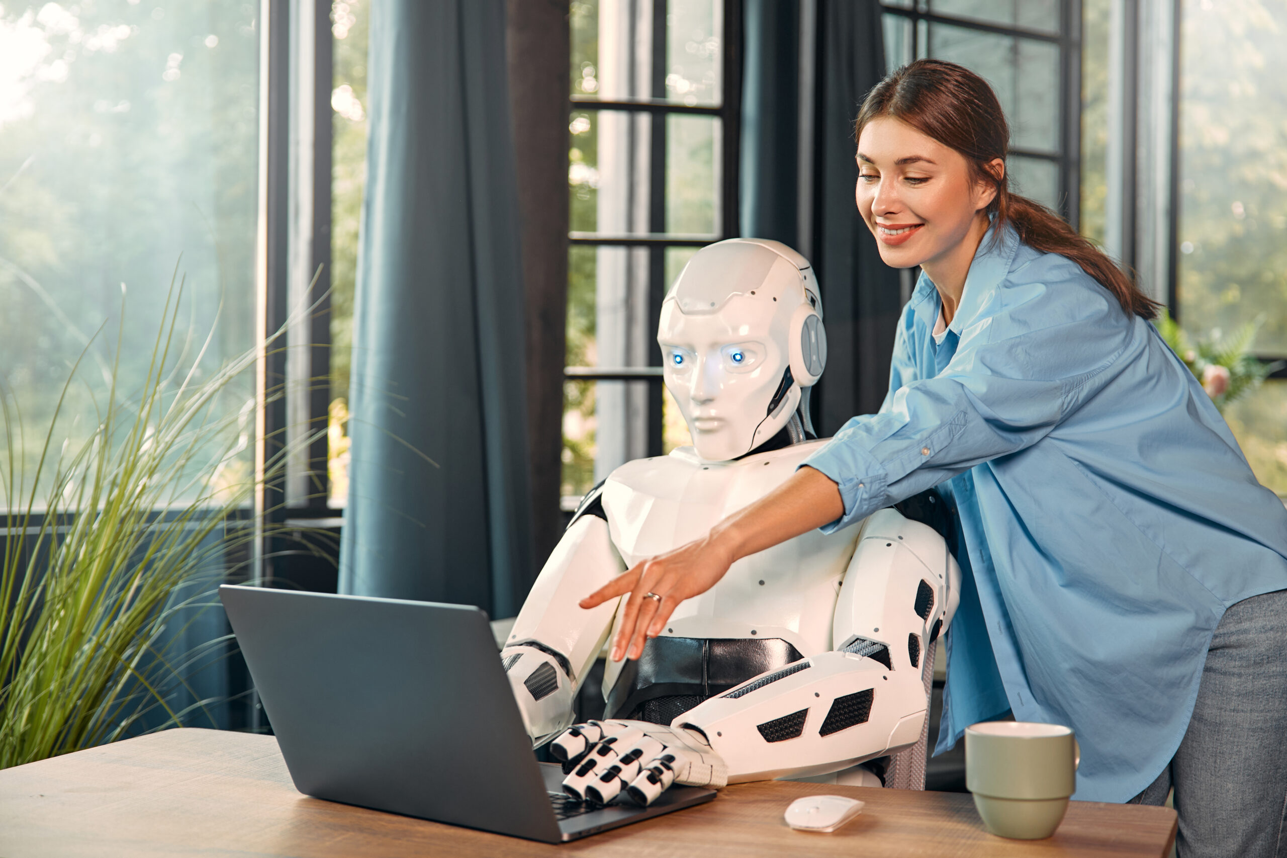 Young woman with a cup of coffee and a humanoid robot working while sitting at a laptop in a modern office. Collaboration between humans and artificial intelligence.