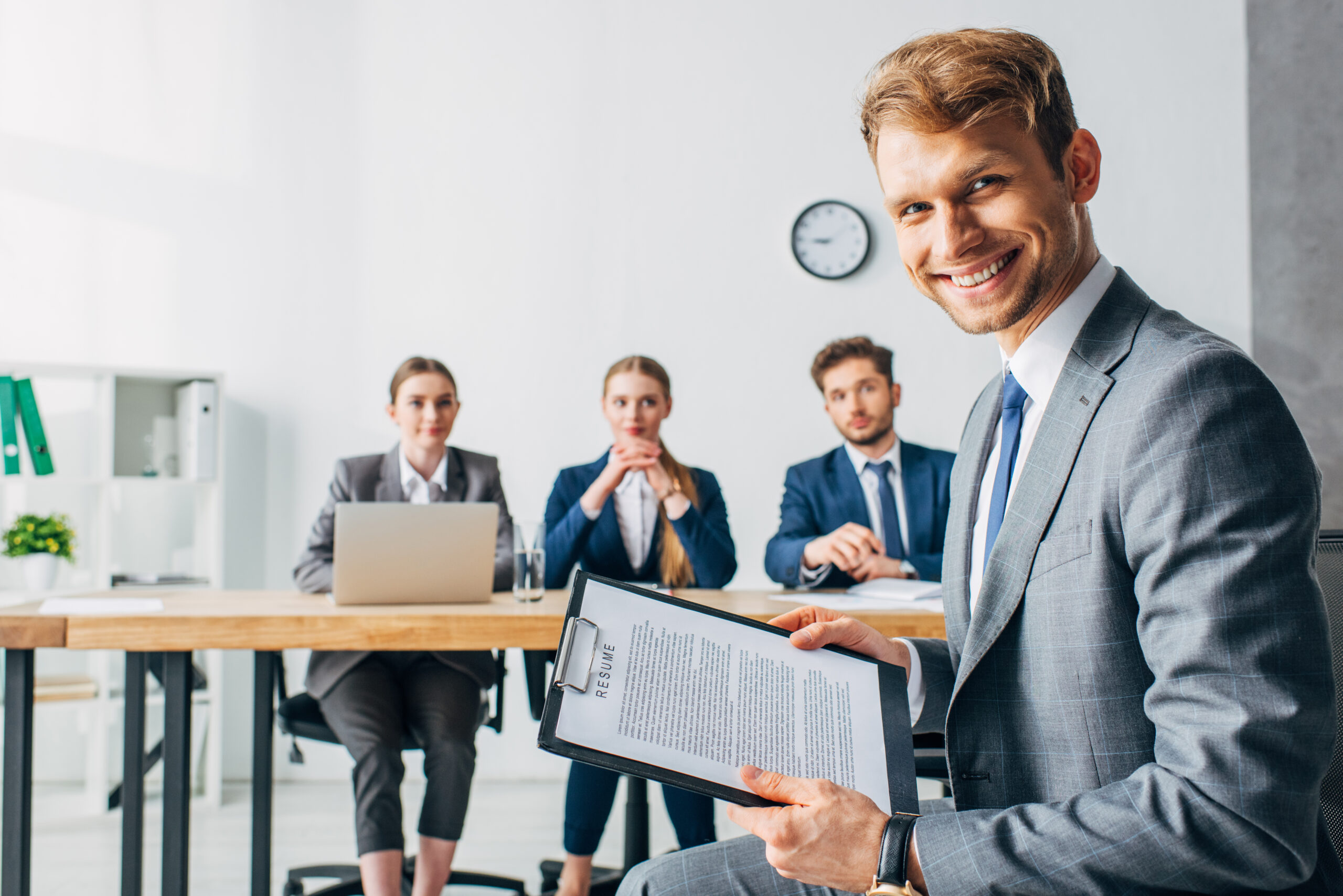 Selective focus of smiling employee with resume looking at camera near recruiters in office Selective focus of smiling employee with resume looking at camera near recruiters in office